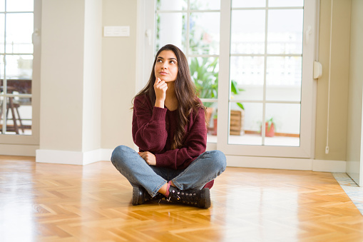 Young beautiful woman sitting on the floor at home with hand on chin thinking about question, pensive expression. Smiling with thoughtful face. Doubt concept.