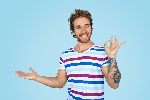 Attractive young male cheerfully smiling and showing OK gesture while demonstrating empty space on blue background