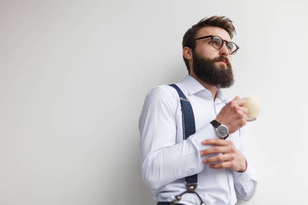 From below of stylish bearded man in eyeglasses and white shirt with suspenders looking away in daylight