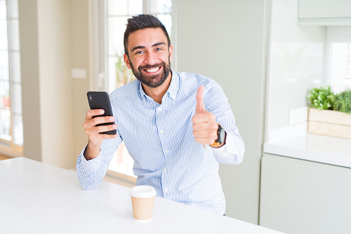 Handsome hispanic business man drinking coffee and using smartphone happy with big smile doing ok sign, thumb up with fingers, excellent sign