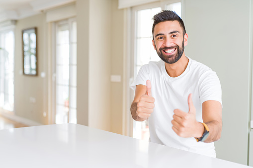 Handsome hispanic man casual white t-shirt at home approving doing positive gesture with hand, thumbs up smiling and happy for success. Looking at the camera, winner gesture.