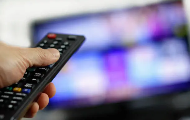 Hand holding remote control with blurred TV set in the background