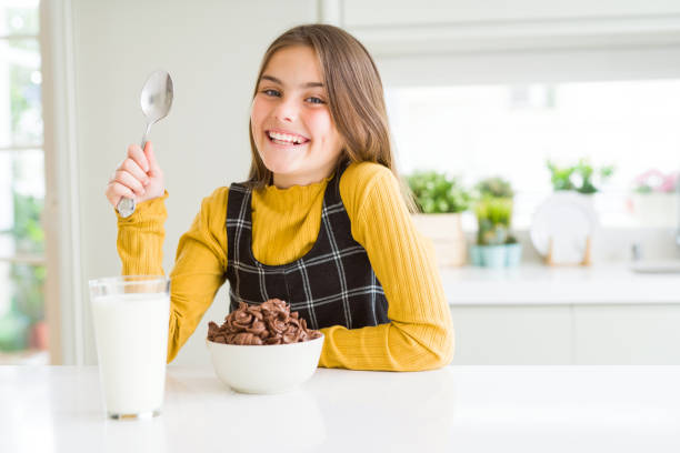 schönes junges mädchen, das schokoladenkergetreide und glas milch zum frühstück mit einem glücklichen gesicht isst und lächelt mit einem selbstbewussten lächeln, das zähne zeigt - toothy smile relaxation white healthcare and medicine stock-fotos und bilder