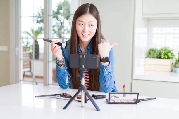 istock Beautiful Asian influencer woman recording make up tutorial using smartphone camera pointing and showing with thumb up to the side with happy face smiling 1134260518
