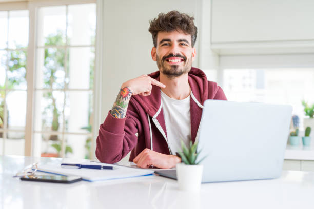 istock Young student man using computer laptop and notebook with surprise face pointing finger to himself 1134262648