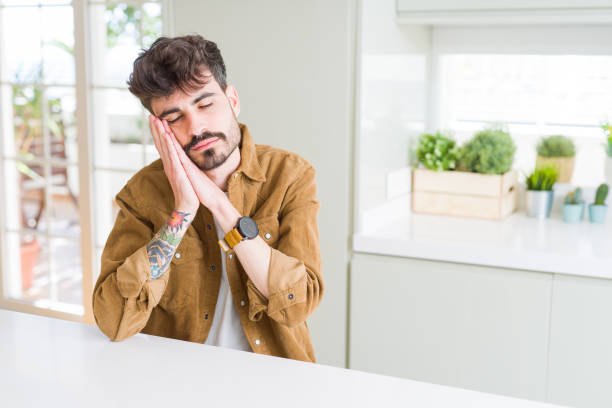 istock Young man wearing casual jacket sitting on white table sleeping tired dreaming and posing with hands together while smiling with closed eyes. 1134262654