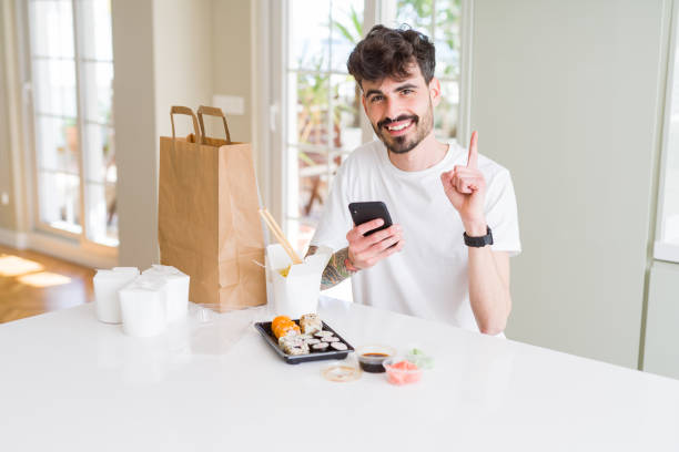 istock Young man eating asian sushi from home delivery and ordering food using smartphone app surprised with an idea or question pointing finger with happy face, number one 1134263379