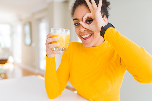 Young african american woman drinking a glass of fresh orange juice with happy face smiling doing ok sign with hand on eye looking through fingers