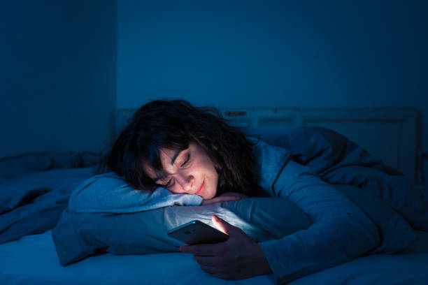 Addicted young woman chatting and surfing on the internet using her smart phone sleepy, bored and tired late at night. Dramatic dark light. In Internet, Mobile addiction and insomnia concept. stock photo