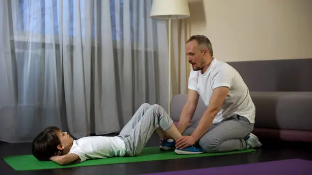Dad holding son legs, training to do crunches, healthy lifestyle, active leisure