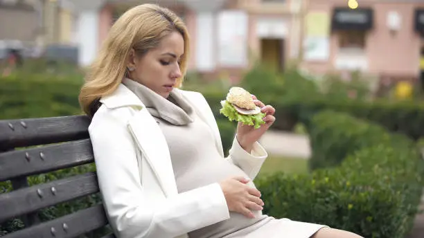 Mother-to-be eating harmful burger sitting park, high fat nutrition, junk food