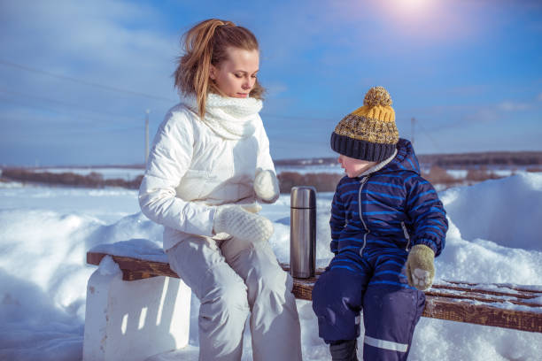 mama und kleiner junge sohn 3-4 jahre alte bank winterstraße, schneeschnee-drifts im hintergrund. thermos mit heißte-suppensuppe trinken, frühstück, mittagessen, erwärmtes kaltes wetter. erholung am kalten wintertag auf dem land. - 3 4 years cute beauty beautiful stock-fotos und bilder