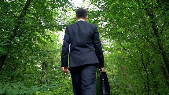 Businessman with briefcase walking in forest, enjoying view, escape from city