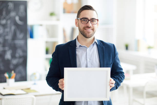 istock Smiling handsome young man in glasses standing in classroom and showing certificate in frame 1134289095