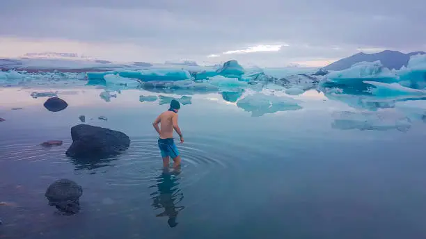 Young man getting into the water of Glacier lagoon. Man wearing only swimming shorts and a hat. Ice bergs drifting in the lagoon. Cold temperatures for ice swimming. Calm surface of the water.