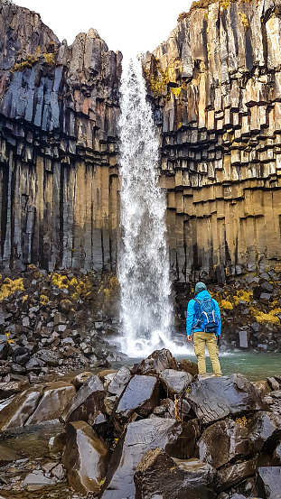 A young man wearing hiking backpack, standing in front of a waterfall. Rocks on the ground look slippery. The water falls from a rock formation, decorated with moss. Svartifoss in Iceland.