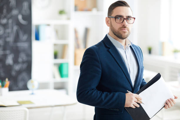 istock Serious confident handsome school teacher with stubble wearing formal jacket and glasses holding clipboard with papers and looking at camera 1134289420
