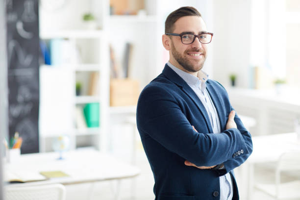 istock Cheerful successful handsome young school teacher with stubble standing in classroom and looking at camera 1134289560
