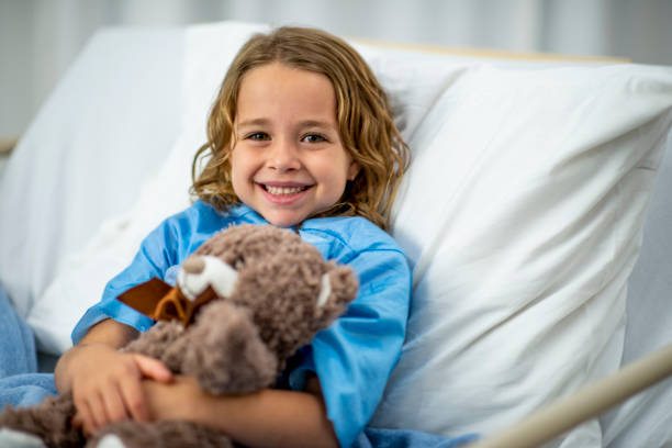 Girl In Hospital Bed A Caucasian girl is indoors in a hospital room. She is smiling at the camera while holding a teddy bear. sick child hospital bed stock pictures, royalty-free photos & images