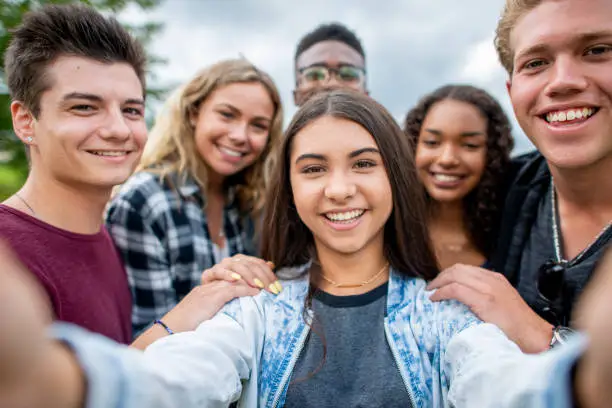 A mixed group of teenaged friends take a selfie together. They have their hands on each others shoulders and show genuine smiles.