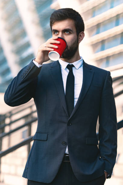 close-up d'un jeune homme d'affaires dans un costume de boire du café à partir d'une tasse de carton rouge en début de matinée près du bureau. - suit necktie close up gray photos et images de collection