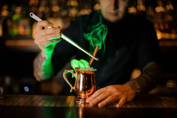 Professional bartender adding to a cocktail in the cooper glass with a dried orange aromatic smoked cinnamon with tweezers on the bar counter