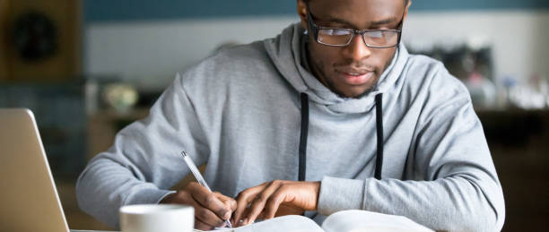 Horizontal photo african student study writing using book and computer Closeup smart millennial african student wear glasses hold pen noting writing down information study use book preparing for university or college test exam, horizontal photo banner for website header exam student university writing stock pictures, royalty-free photos & images
