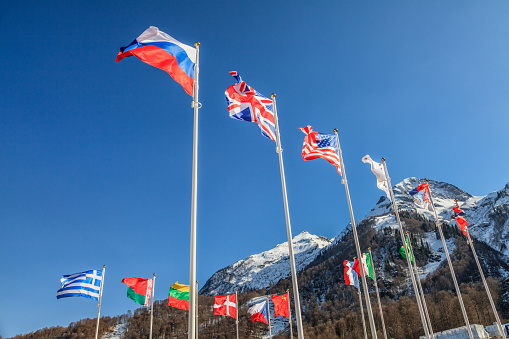 National flags of Russia, UK, USA, Greece, other countries wave in wind as friendship symbol on mountain peaks and blue sky background. Various fluttering national flags on flagpoles. Sochi, Russia.