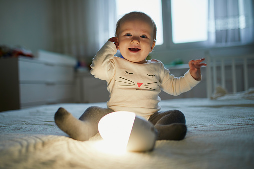 Adorable baby girl playing with bedside lamp in nursery. Happy kid sitting on bed with nightlight. Little child at home in the evening before sleep