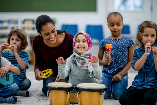 A multi-ethnic group of young school children are indoors in their classroom. Their teacher is watching them playing instruments together. The instruments include drums, maracas, and a guitar.