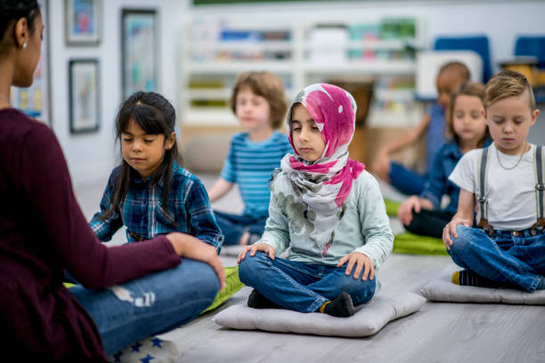 Yoga Class A multi-ethnic group of young school children are indoors in their classroom. They are sitting on pillows and doing yoga together. They are sitting with their hands in their lap. mindfulness kids stock pictures, royalty-free photos & images