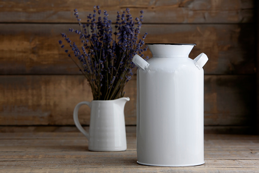 White metal milk jug next to a vase of lavender on a wooden background. Mockup is ready for your design or logo to be added.