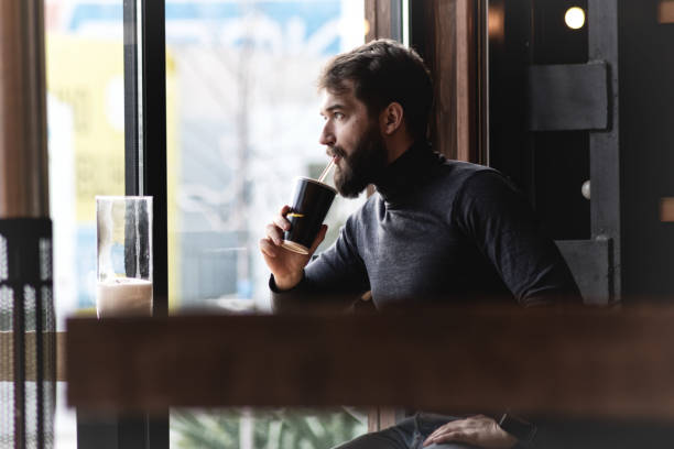 istock handsome bearded guy drinks a drink from a straw near the window in a cafe.  attractive young stylish man sitting in a cafe and looking out the window. Business, freelance 1134348123