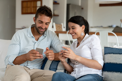 Happy couple relaxing at home looking at social media on their cell phones and smiling - lifestyle concepts