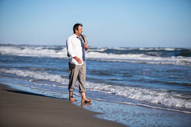 Middle-aged corporate man with loose white shirt and tie and rolled-up pants calmly walking barefoot on a beach with the coat on his shoulder stock photo