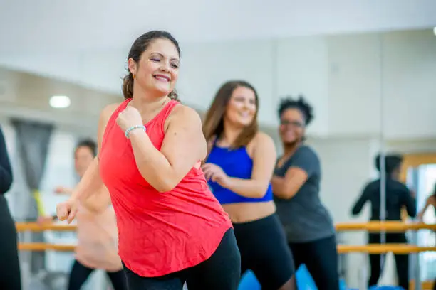 A woman smiles as she dances her way to fitness. Her hair is flowing to the side and her arms are outstretched. She is wearing workout clothes.