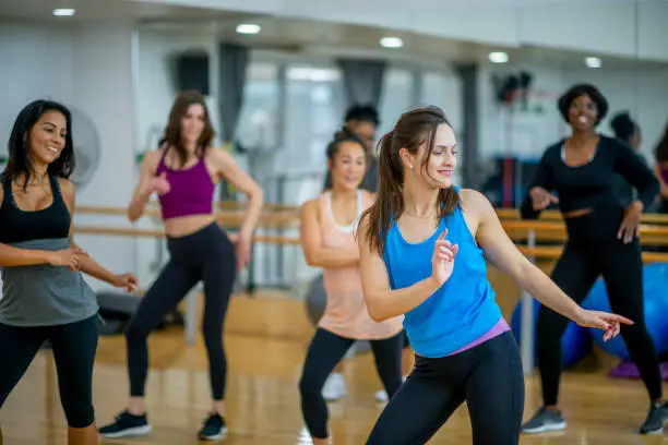 A diverse group of women dances in their fitness class. They are all laughing while their hands are stretched out to the side.