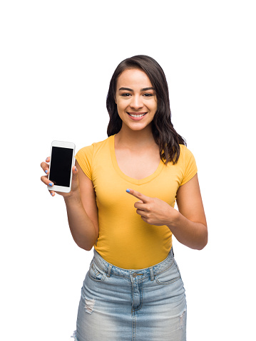 Latin young woman with brown hair pointing at a smart phone smiling looking at camera in a mid shot isolated on a white background.