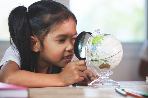 Cute asian child girl use magnifier to look and study at the globe in classroom