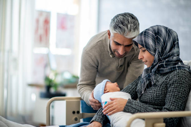 New Mother And Father A mother and father are indoors in a hospital room. The mother is wearing a head scarf, and she is holding her newborn baby boy. The father is touching his son and watching him sleep. muslim mother and baby stock pictures, royalty-free photos & images