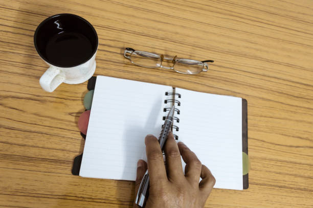 istock Businessman writing in his diary. Open notebook with blank pages next to cup of coffee and eyeglasses on wooden table. Business still life concept with office stuff on table. Top view with copy space. 1134407164