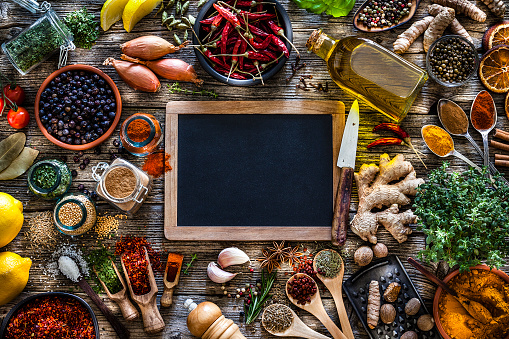 Top view of a rustic wood kitchen table with a large group of multi colored spices and herbs placed all around a blank blackboard leaving useful copy space for text and/or logo. Spices and herb included are clove, turmeric, star anise, bay leaf, cinnamon, curry powder, ginger, nutmeg, peppercorns, cinnamon, salt, chili pepper, cardamom, dried oregano, basil, parsley, lemon, rosemary, garlic, onion and dried orange slices. An olive oil bottle is included in the composition as well as a closed cookbook. DSRL studio photo taken with Canon EOS 5D Mk II and Canon EF 70-200mm f/2.8L IS II USM Telephoto Zoom Lens