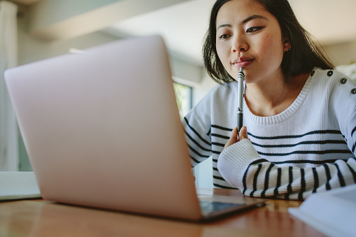 Asian woman studying at home using laptop on the table. Young female using laptop holding a pen in hand.