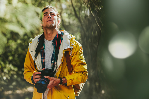 Portrait of a man wearing jacket and backpack walking in a forest holding a dslr camera. Man exploring a forest capturing the beauty in a digital camera.