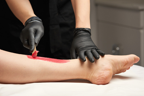 woman with barefoot leg, beautician removing hair with red wax.
