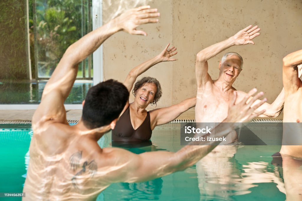 Males and females exercising with arms raised Smiling instructor with senior men and woman during aqua aerobics. Males and females are exercising with arms raised in swimming pool. They are at nursing home. Exercising Stock Photo