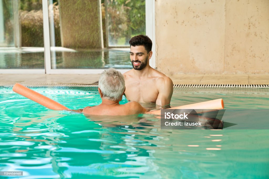 Smiling instructor assisting man with noodle float Smiling shirtless men exercising in swimming pool. Instructor is assisting senior male with noodle float. They are at nursing home. Rear View Stock Photo