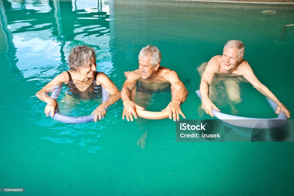 Males and female swimming with noodle floats High angle view of senior woman and men with noodle floats. Retired males and female are swimming in pool. They are exercising at nursing home. Senior Adult Stock Photo