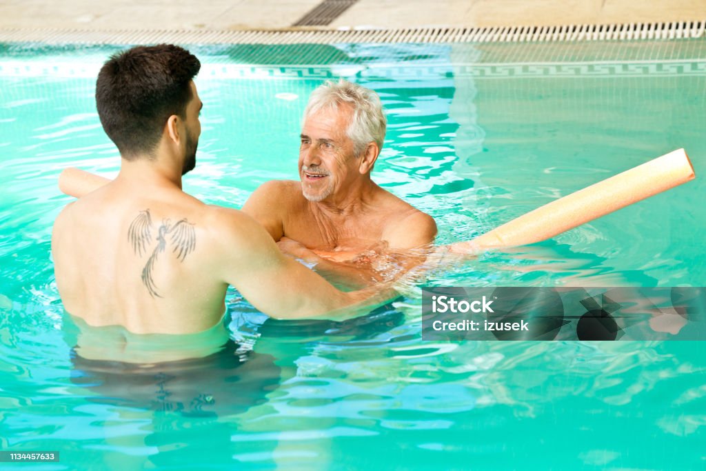 Instructor assisting senior man with noodle float Instructor assisting senior male with noodle float. Shirtless men are exercising in swimming pool. They are at nursing home. Water Aerobics Stock Photo