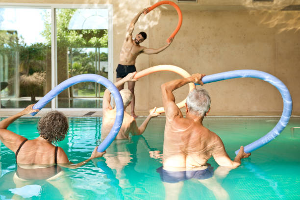 Males and females exercising in swimming pool Instructor exercising with senior men and woman during aqua aerobics. Males and females are holding noodle floats in swimming pool. They are at nursing home. community health center stock pictures, royalty-free photos & images
